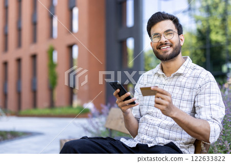Portrait of a young Indian man holding a credit card and a mobile phone, sitting on a bench outside a building and looking at the camera. Portrait of a young Indian man holding a credit card and a mobile phone, sitting on a bench outside a building and looking at the camera. 122633822