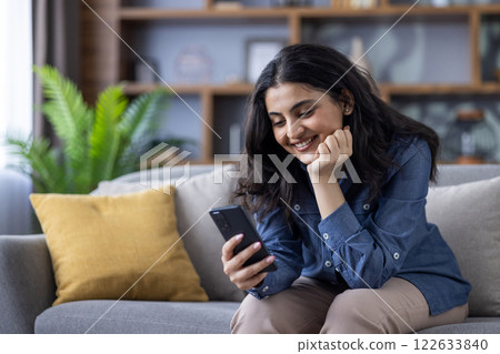 A dreamy and pensive young Indian girl is sitting on the sofa at home, resting her head on her hand and smilingly looking at the mobile phone screen. 122633840