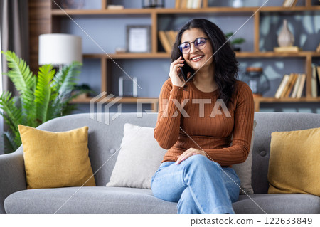 Smiling young Indian woman sitting relaxed on sofa at home and talking on mobile phone. 122633849