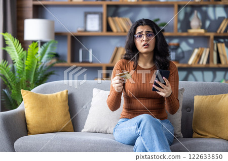 Portrait of upset young Indian woman in glasses sitting on sofa at home, holding credit card and phone, looking worriedly at camera. 122633850