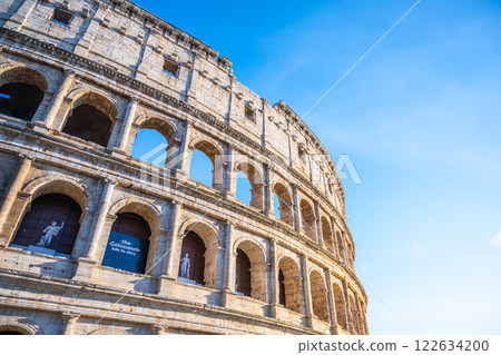 Visitors admire the grand structure of the Colosseum in Rome, Italy, showcasing its impressive ancient architecture against a clear blue sky, highlighting its historical significance and beauty. 122634200