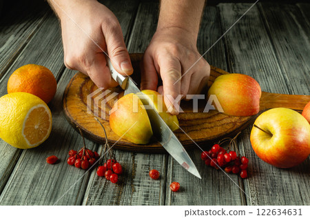 Hands deftly slice a ripe apple on a rustic wooden board surrounded by vibrant fruits. The bright colors of lemons and berries enhance the appetizing display, inviting a healthy feast 122634631