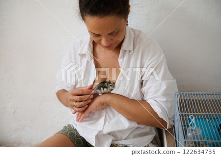 Woman holding a hamster inside her shirt while sitting by a pet's cage 122634735