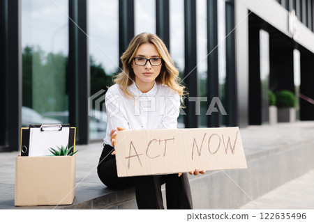 Woman sitting outdoor with poster Act Now next to box of stuff . Beautiful female protester at strike against unemployment 122635496