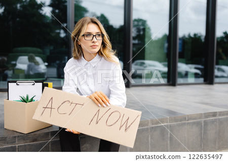 Woman sitting outdoor with poster Act Now next to box of stuff . Beautiful female protester at strike against unemployment 122635497