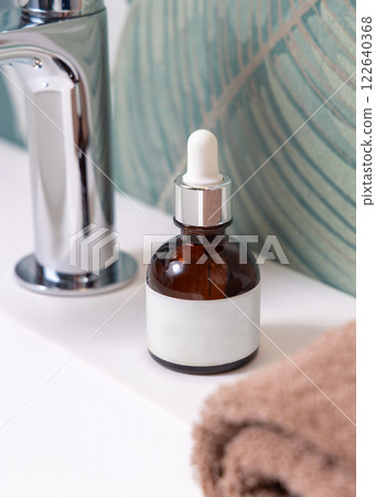 Dropper bottle with label near brown towels on basin in tropical bathroom close up, cosmetic mockup 122640368
