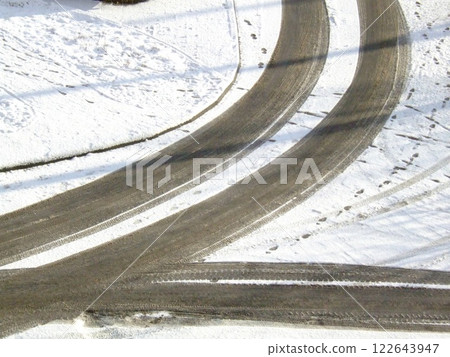 Tire tracks in the snow create winding patterns on a road. The scene captures a peaceful suburban area blanketed in white, with gentle curves and a serene atmosphere. Tire tracks in the snow create winding patterns on a road. The scene captures a peaceful suburban area blanketed in white, with gentle curves and a serene atmosphere. 122643947