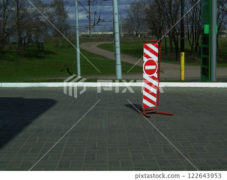 A vibrant caution sign indicating no entry stands prominently on a paved area. The background features a winding path and lush trees under a partly cloudy sky, suggesting early afternoon. 122643953