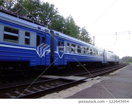 A blue passenger train rests at a quiet rural station surrounded by trees in the early morning light. The platform is empty, showcasing the serene atmosphere of the moment. A blue passenger train rests at a quiet rural station surrounded by trees in the early morning light. The platform is empty, showcasing the serene atmosphere of the moment. 122643954