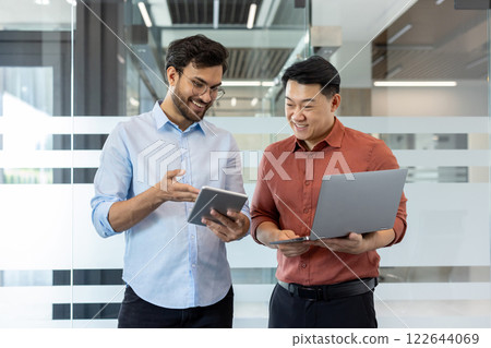 Two programmer colleagues in casual clothes discussing a new software project, men standing by the window with a laptop, smiling, talking. Two programmer colleagues in casual clothes discussing a new software project, men standing by the window with a laptop, smiling, talking. 122644069