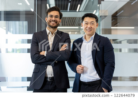 Two confident businessmen standing in modern office. One with arms crossed and other with hands in pockets. They express teamwork, professionalism, and success in business environment. 122644079