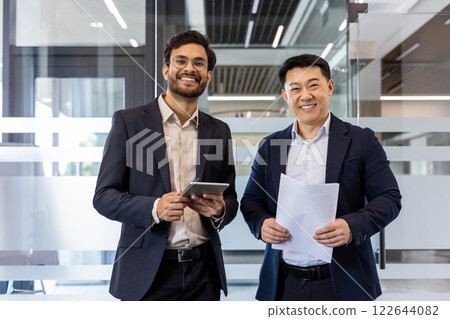 Smiling businessmen standing in modern office showcasing teamwork and professionalism. One holding tablet, other documents, reflecting success and collaboration in business environment. Smiling businessmen standing in modern office showcasing teamwork and professionalism. One holding tablet, other documents, reflecting success and collaboration in business environment. 122644082