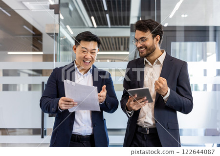 Two business professionals sharing moment of success in office. One holding paperwork, other with tablet. Expressions of joy and achievement visible on faces. Teamwork and collaboration 122644087