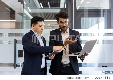 Two businessmen engaged in discussion holding laptop and tablet in modern office. Displaying teamwork, communication in professional business environment. Emphasis on collaboration, technology Two businessmen engaged in discussion holding laptop and tablet in modern office. Displaying teamwork, communication in professional business environment. Emphasis on collaboration, technology 122644114