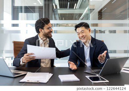 Two cheerful professionals are sitting at a modern office table, engaged a lively conversation. The environment suggests a positive collaboration, with documents, laptops, indicating work activities Two cheerful professionals are sitting at a modern office table, engaged a lively conversation. The environment suggests a positive collaboration, with documents, laptops, indicating work activities 122644124