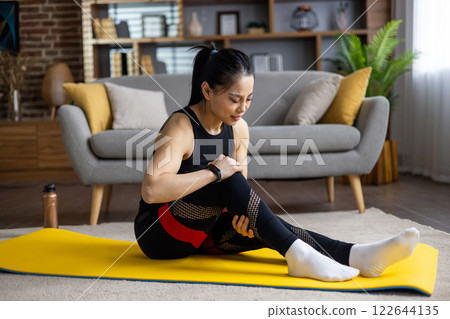 Asian woman injured while doing fitness and yoga at home, woman strained leg muscle, massaging, muscle pain, sitting on sports mat in living room in apartment. 122644135