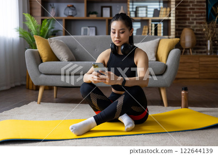 Asian woman in workout attire using smartphone and headphones during home exercise session on yoga mat. Engaged in fitness activity, reflecting contemporary lifestyle and mindfulness practices. 122644139