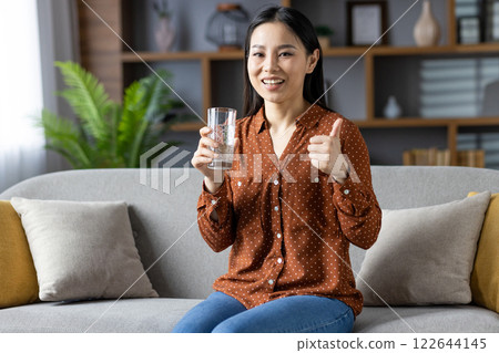 Asian woman smiling on sofa holding glass of water while giving thumbs up. Wearing casual brown polka dot shirt, jeans, radiating positivity, relaxation, well-being, hydration, healthy lifestyle. 122644145