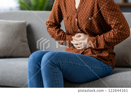 Young woman with stomach pain sitting on sofa, holding abdomen, displaying discomfort. She wears brown polka-dot shirt, blue jeans, conveying concepts of health, pain, and discomfort. 122644161