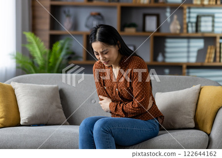 Asian woman on sofa holds stomach with discomfort. Suffering from stomachache or digestive issues. Casual indoor setting at home conveys pain and discomfort. Asian woman on sofa holds stomach with discomfort. Suffering from stomachache or digestive issues. Casual indoor setting at home conveys pain and discomfort. 122644162