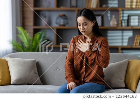 Asian woman experiencing chest discomfort sitting on sofa. Image conveys health concern or stress. Cozy living room setting enhances focus on emotional expression and wellness. Concept of difficulty. 122644168