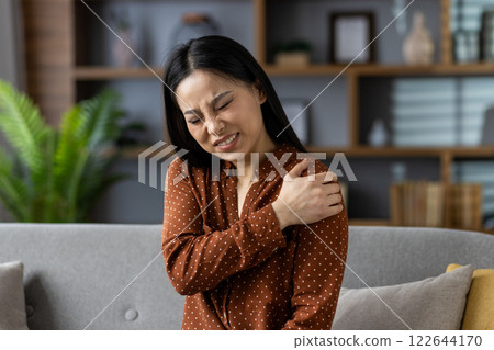 Asian woman sitting on sofa with shoulder pain, seeking relief. Expression shows discomfort, wearing polka dot blouse in modern living room setting. Concept of physical discomfort. 122644170