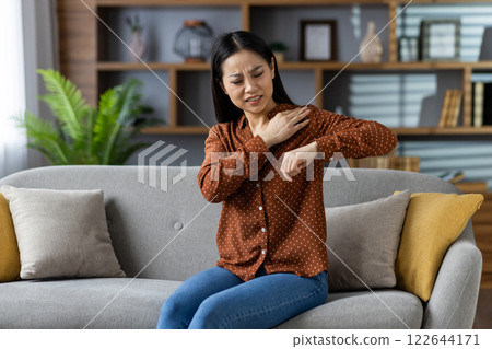 Asian woman experiencing shoulder pain while sitting on sofa in home interior. She touches sore shoulder, wearing casual polka dot blouse, expressing discomfort, conveying health and wellness concern Asian woman experiencing shoulder pain while sitting on sofa in home interior. She touches sore shoulder, wearing casual polka dot blouse, expressing discomfort, conveying health and wellness concern 122644171