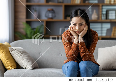 Asian woman sits on gray sofa feeling thoughtful, alone. Cozy living room with modern decor, cushions. Emotions conveyed include contemplation, solitude, reflection, mindfulness. Asian woman sits on gray sofa feeling thoughtful, alone. Cozy living room with modern decor, cushions. Emotions conveyed include contemplation, solitude, reflection, mindfulness. 122644182