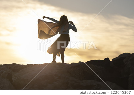 A woman is standing on a rocky hillside, with the sun setting in the background A woman is standing on a rocky hillside, with the sun setting in the background 122645307