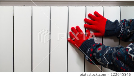 Hands in gloves on a radiator. Central heating radiator at home. A child in woolen gloves warms his hands near the heater Hands in gloves on a radiator. Central heating radiator at home. A child in woolen gloves warms his hands near the heater 122645546