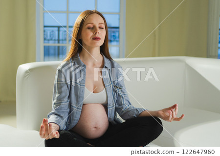 Pregnancy prenatal breathing. Pregnant woman with big belly sitting in lotus pose on sofa at home. Pregnant girl practicing yoga mindfulness meditation. Relaxation self care during pregnancy Pregnancy prenatal breathing. Pregnant woman with big belly sitting in lotus pose on sofa at home. Pregnant girl practicing yoga mindfulness meditation. Relaxation self care during pregnancy 122647960
