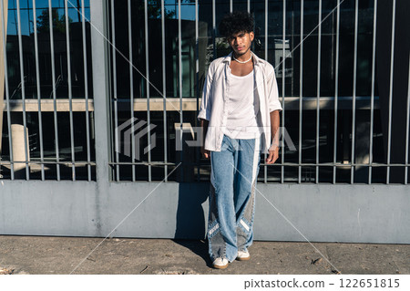 A handsome young Latino man leaning on a large fence at the entrance of a building with a serious expression 122651815