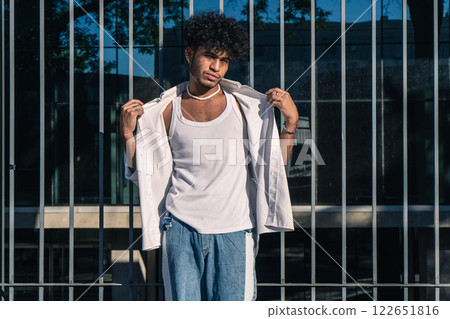 A handsome young Latino man leaning on a large fence at the entrance of a building with a serious expression 122651816