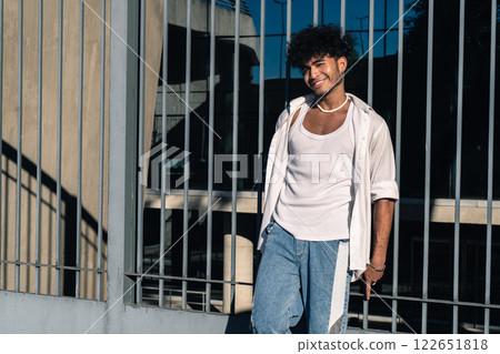 A handsome young Latino man leaning on a large fence at the entrance of a building with a friendly and happy expression 122651818