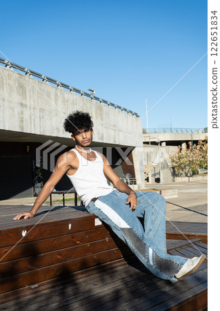 Young Latino man sitting on a wooden platform in a square Young Latino man sitting on a wooden platform in a square 122651834