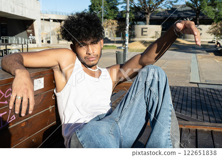 Young Latino man sitting on a wooden platform in a square Young Latino man sitting on a wooden platform in a square 122651838