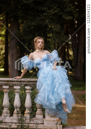 Romantic Woman in a Light Blue Gown Posing on a stone balustrade in a picturesque garden. 122654121
