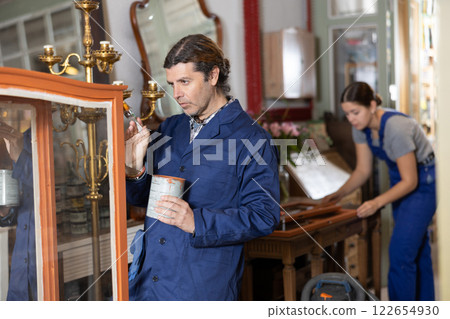 Skilled middle-aged male furniture repair specialist in uniform restoring and painting cupboard furniture during workday in workshop 122654930