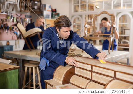 Skilled middle-aged male furniture repair specialist in uniform restoring Victorian dresser during workday in workshop Skilled middle-aged male furniture repair specialist in uniform restoring Victorian dresser during workday in workshop 122654964