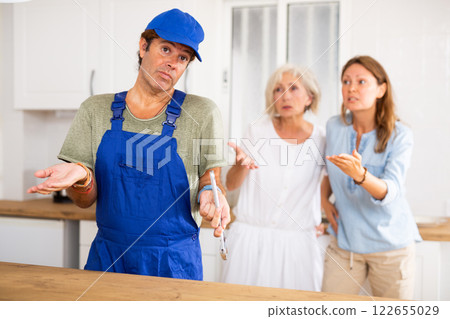 Inattentive plumber in uniform spreading his arms to the sides while being scolded by two women in the kitchen Inattentive plumber in uniform spreading his arms to the sides while being scolded by two women in the kitchen 122655029
