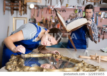 Girl restorer carefully covers carved wooden frame of antique wall mirror with thin layer of varnish Girl restorer carefully covers carved wooden frame of antique wall mirror with thin layer of varnish 122655055