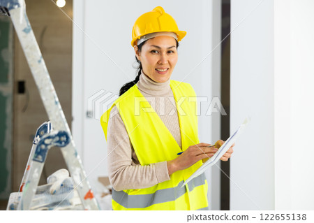 Woman foreman in a protective helmet and yellow vest checks the completed construction work on drawing Woman foreman in a protective helmet and yellow vest checks the completed construction work on drawing 122655138