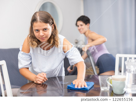 Girl cleaning surface on table while girlfriend cleaning floor with vacuum cleaner 122655156