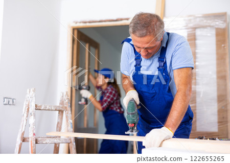 Concentrated male builder in uniform using a jigsaw machine on wooden plank at the apartment Concentrated male builder in uniform using a jigsaw machine on wooden plank at the apartment 122655265