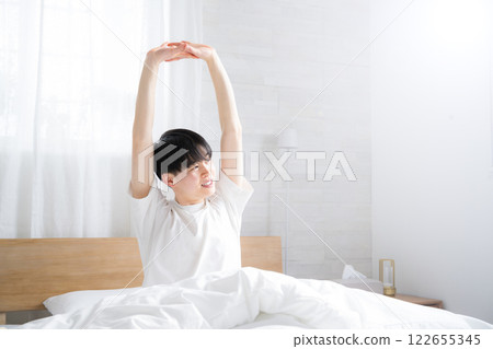 一個年輕人在床上醒來，沐浴著早晨的陽光，伸展著雙臂 122655345