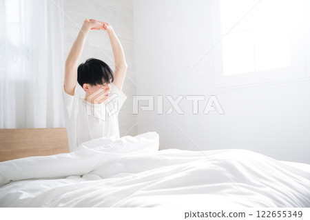 A young man waking up in bed bathed in the morning sun stretching his arms with copy space on the right and eyes closed 122655349