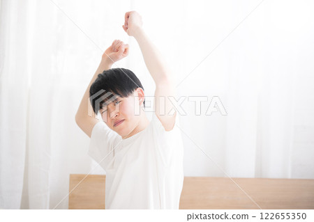 A young man waking up in bed bathed in the morning sun stretching his arms with copy space on the right and eyes closed A young man waking up in bed bathed in the morning sun stretching his arms with copy space on the right and eyes closed 122655350