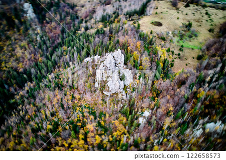Dynamic aerial view of rocks highlights rugged, rocky outcrop amidst vibrant, autumnal forest. Surrounding landscape, with mix of dense trees and open fields, creates dynamic and colorful scene. 122658573