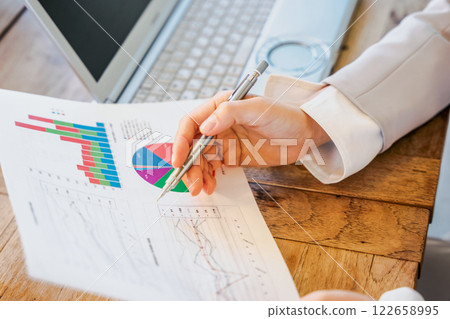 A woman's hands pointing at a paper document with a pen in front of a computer 122658995