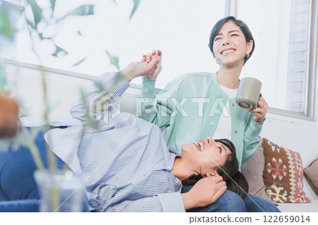 Two same-sex partners sitting on a sofa, holding hands and resting their heads on their partner's laps, exchanging physical contact and confirming their affection 122659014